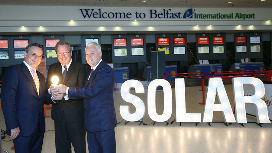 Three men holding a lit lightbulb at Belfast Airport, a lit up sign saying 'Solar' in the background