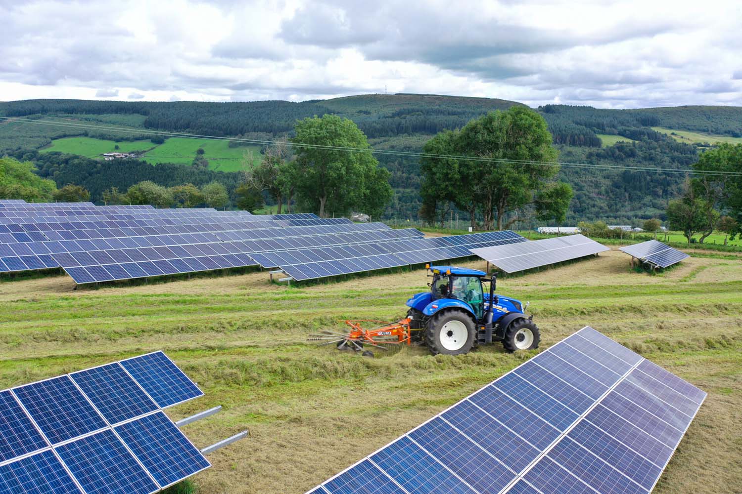 A blue tractor mows grass between rows of solar panels at a Lightsource bp solar project in the UK, part of their UK Projects portfolio, set against rolling green hills under a cloudy sky.