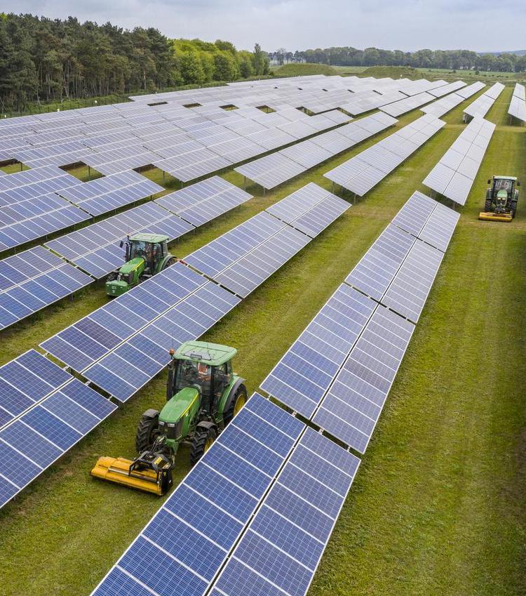 Wide aerial view of solar panels spanning a large grassy area, with tractors working between rows of panels.