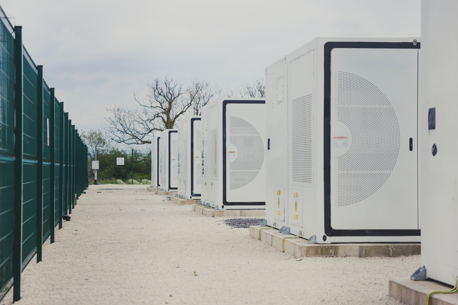 Row of white, industrial-scale Battery Energy Storage System (BESS) units, enclosed within a green perimeter fence at a utility-scale solar farm site, demonstrating energy storage infrastructure and security.