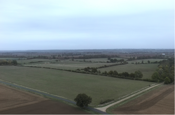 Aerial view of vast, flat farmland and green fields divided by trees and hedgerows under an overcast, cloudy sky, depicting a rural landscape or potential site location area.

