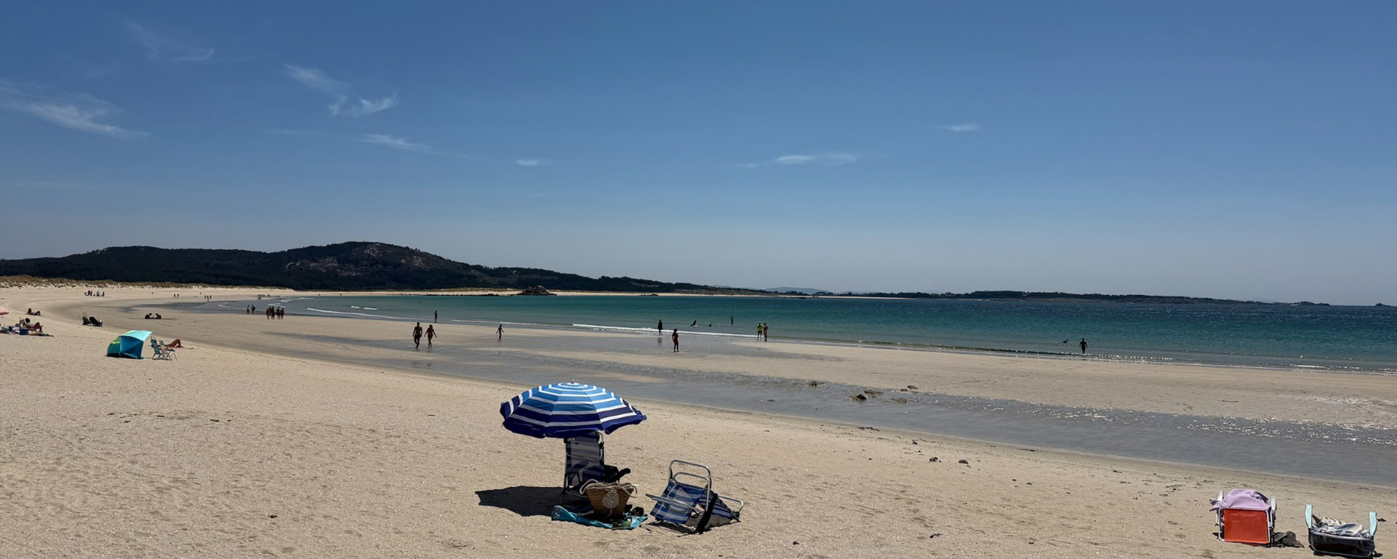 A sunny sandy beach with a blue and white striped umbrella shading a person, scattered beach chairs, calm ocean, and distant hills under a clear blue sky.
