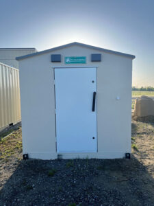 A small, white, prefabricated tornado shelter with a closed door, located outdoors on a gravel surface, with a sign above the door that reads "Pro Storm Shelters."