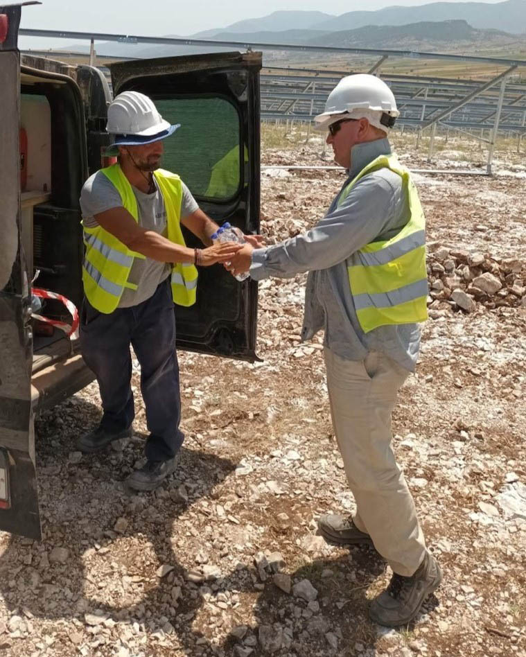 Two construction workers in PPE on a solar site, handing bottles of water between them. Solar panels in the background behind them.