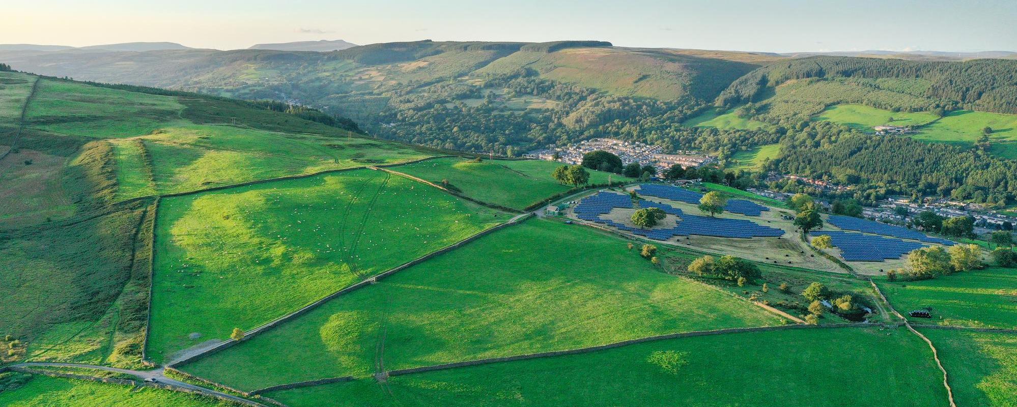 Aerial view of a small solar farm nestled in green rolling hills with a village nearby and forested mountains in the background under a soft, late afternoon light.