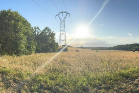 A sunlit rural field with a transmission tower standing against a blue sky, bordered by trees and rolling hills.