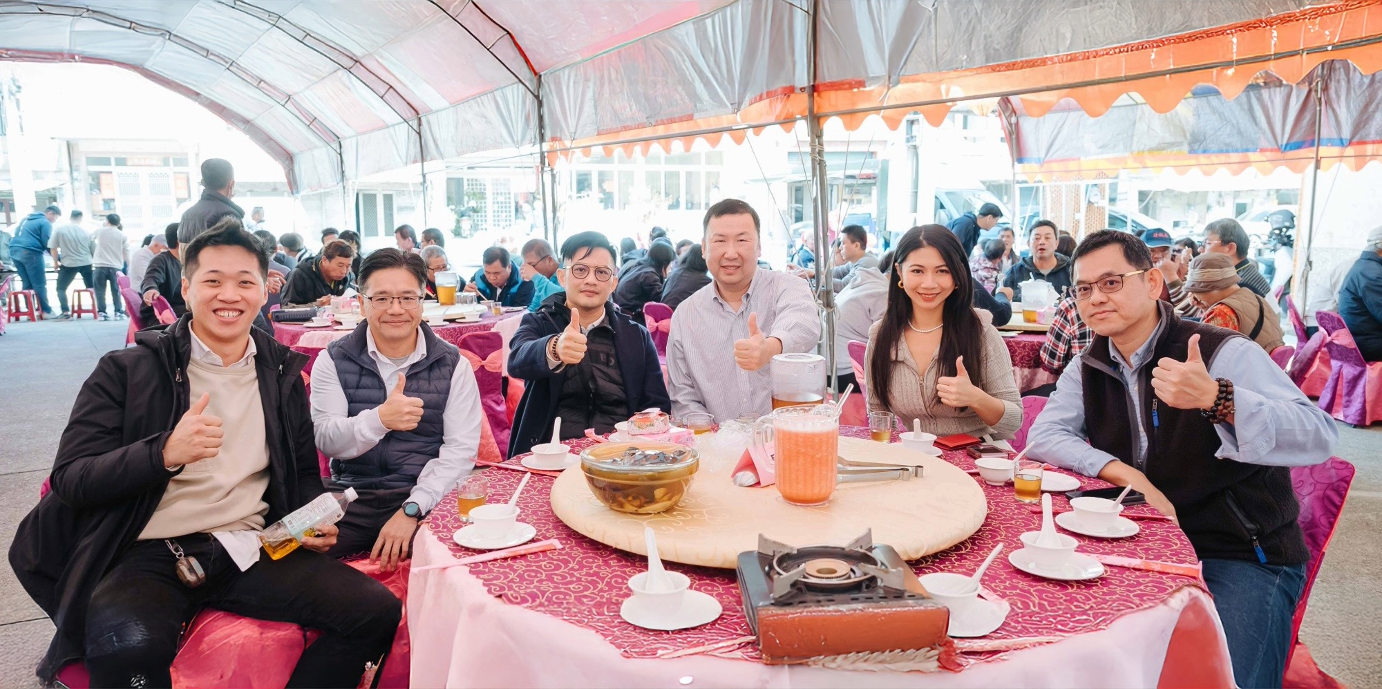 一群人坐在遮棚下圓桌周圍，用紅色桌布覆蓋，豎起大拇指比讚。